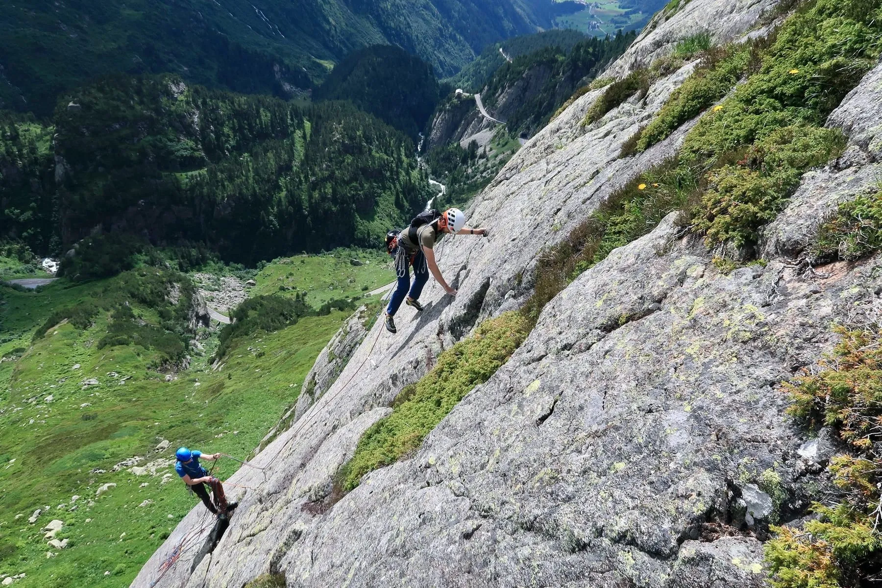 Zwei Kletterer mit Helmen klettern an einer senkrechten Felswand in einer grünen Berglandschaft.